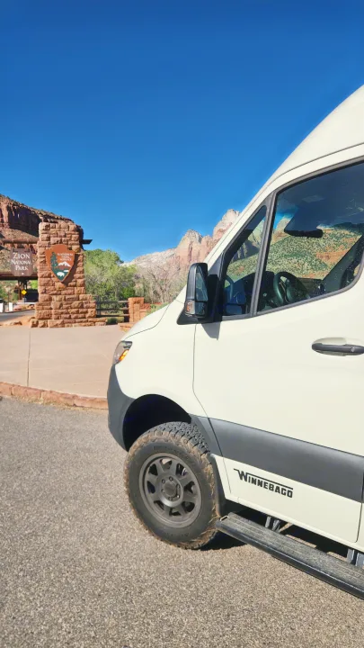 Adventure begins! This white Winnebago RV rental is parked at Zion National Park's stone entrance sign. Majestic red rocks in view.. Winnebago Revel 2024