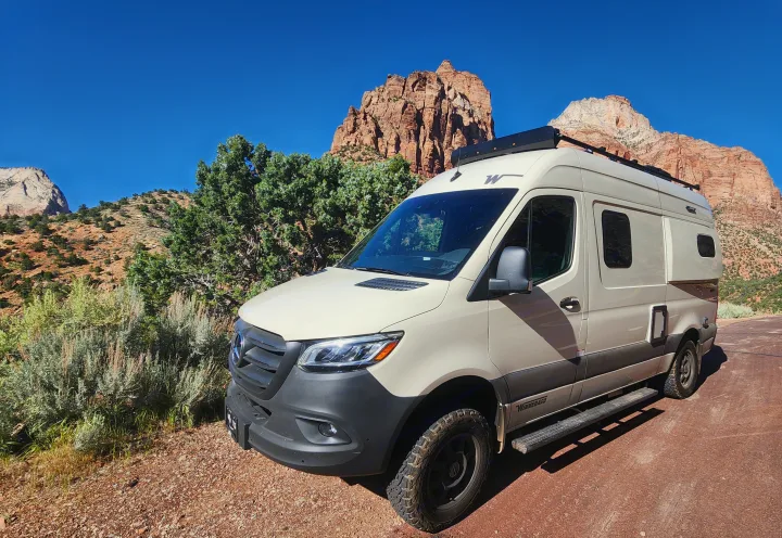 A cream-colored Winnebago RV rental van is parked on a red dirt road amidst towering red rock mountains under a blue sky.. Winnebago Revel 2024