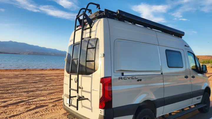 RV rental Winnebago Revel camper van by a lake with mountains, showing its rear ladder, roof rack, and awning.. Winnebago Revel 2024