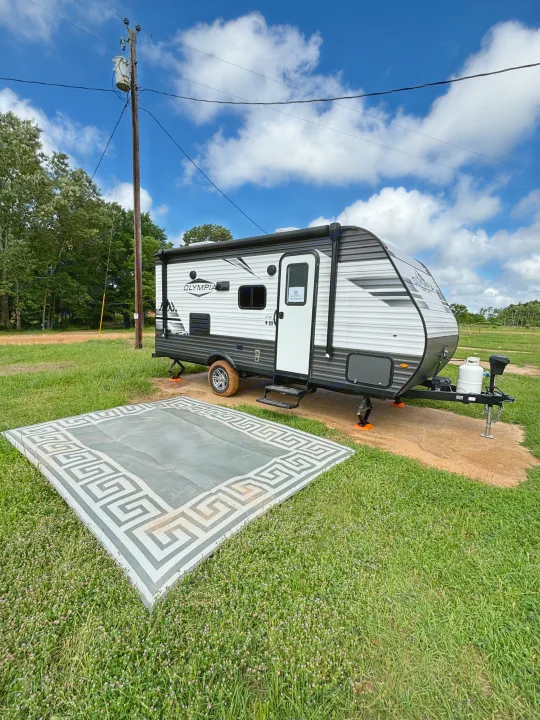 An Olympia Sport RV rental is set up at a rural campsite, with an ornate outdoor rug on grass.. Olympia Sport 19BH 2022
