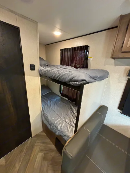 This RV rental's sleeping area features two compact bunk beds with grey bedding, dark curtains, and a seating area.. Olympia Sport 19BH 2022