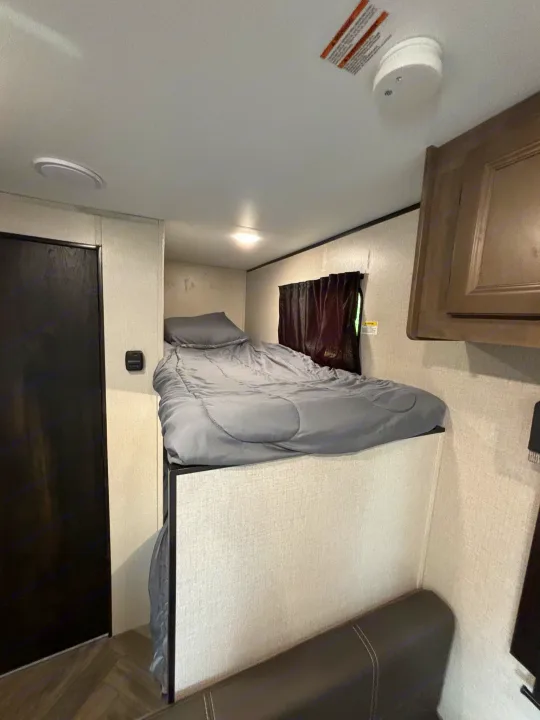 An RV rental interior featuring a cozy bunk bed with grey bedding, window with dark curtain, ceiling light, and smoke detector.. Olympia Sport 19BH 2022