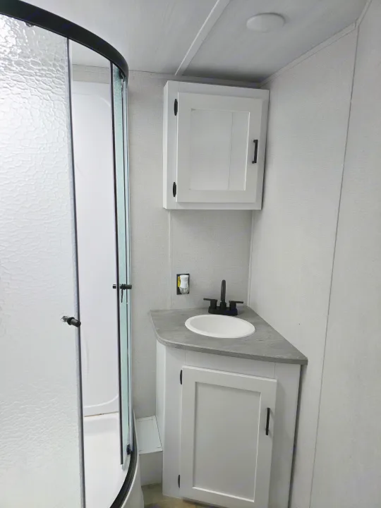 Compact RV rental bathroom features corner vanity, sink, black faucet, wall cabinet, and textured glass shower.. Keystone RV Hideout 2023
