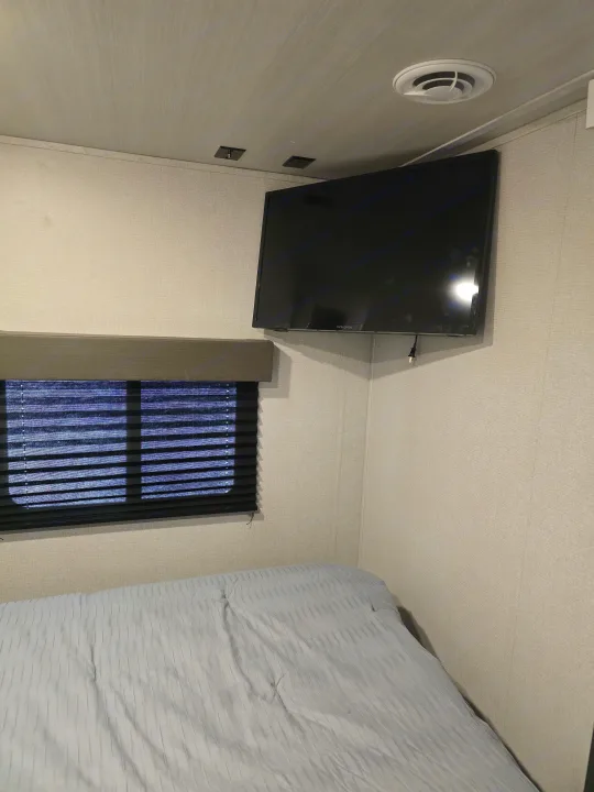 Inside an **RV rental**: a compact bedroom featuring a light blue striped bed, wall-mounted TV, and blinds on the window.. Keystone RV Hideout 2023