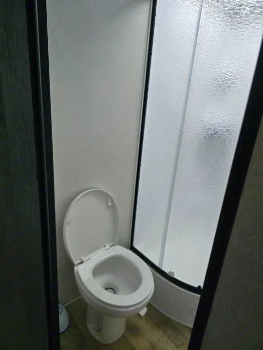 View of a compact RV rental bathroom featuring a white toilet with an open lid and a frosted glass shower enclosure.. Keystone RV Hideout 2023