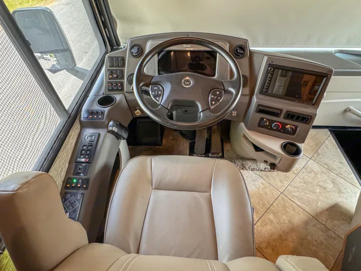 RV rental cockpit: spacious interior with a tan driver's seat, steering wheel, multi-screen dashboard, and various controls.. Winnebago Forza 2019