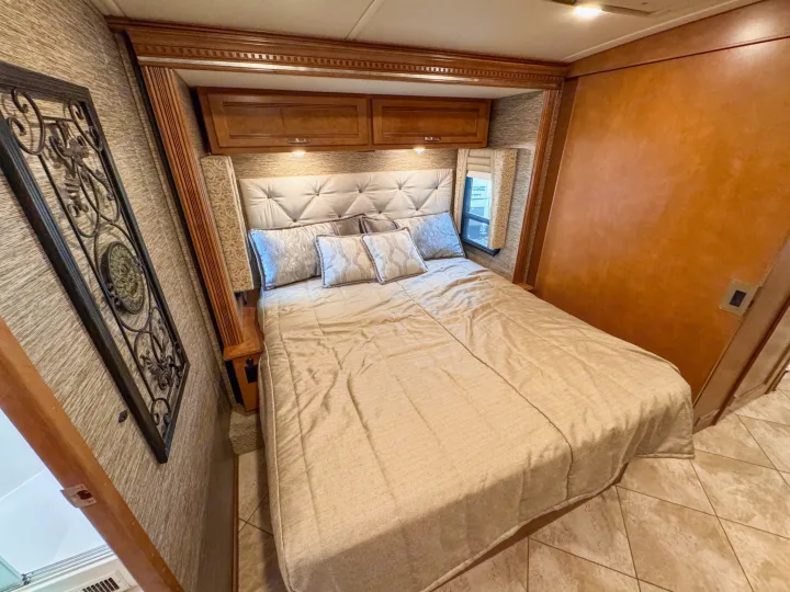 Cozy RV rental bedroom featuring a large bed, tufted headboard, and overhead storage. Ready for comfortable travel.. Winnebago Forza 2019