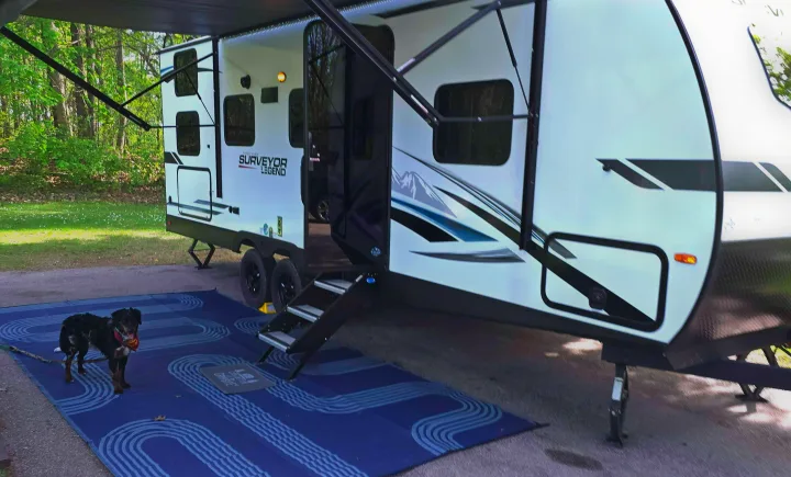 White RV rental travel trailer set up at a campsite with awning, steps, a blue mat, and a dog enjoying the space.. Forest River Surveyor Legend 2023