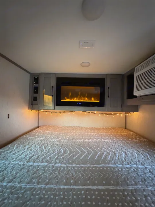 Cozy RV rental bedroom interior with a patterned bed, illuminated by string lights and an electric fireplace, plus an AC unit.. Dutchmen Other 2023
