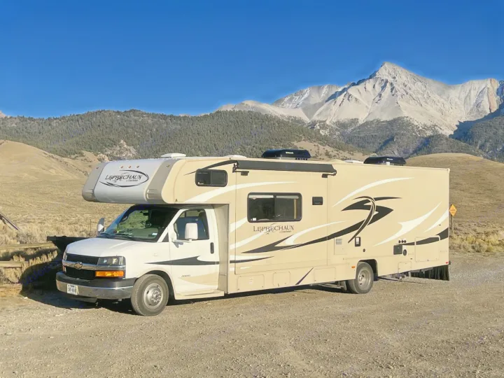 Tan rv rental on gravel, with clear blue sky and majestic mountains in the background. Ideal for scenic nature travel.. Coachman Leprechaun 2014