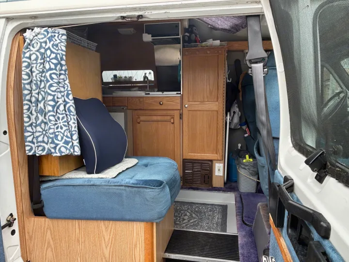This RV rental shows a compact interior: blue cushioned seating, wood cabinets, and a small kitchen with a sink and microwave.. Roadtrek 190 Popular 1997