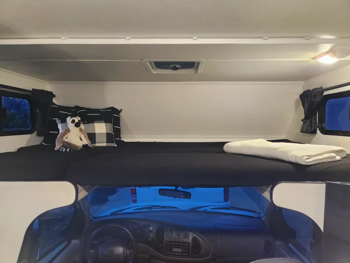 **RV rental** interior: A plush, dark-colored **cab-over bunk** bed with pillows, a lemur toy, and a white blanket. The vehicle's **dashboard** is visible below.. Four Winds Majestic 2008
