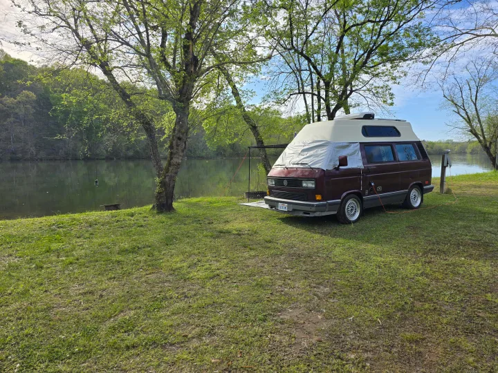 Relaxing by the river: a maroon camper van, hooked up at a scenic spot, perfect for your next RV rental trip.. Volkswagen Westfalia 1990