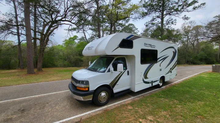 This white Freedom Elite rv rental motorhome is perfectly parked on an asphalt road in a serene wooded park.. Thor 22HE 2019