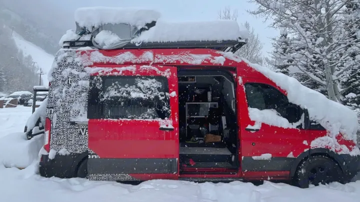 A red camper van, an RV rental, covered in snow in a mountain setting. Its side door is open, revealing the interior living space.. Ford Transit 2016