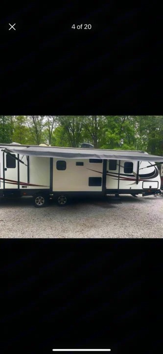 Side view of a white travel trailer RV with an extended awning, parked on a gravel lot under trees, perfect for rv rental.. Heartland RVs Sundance XLT 2016