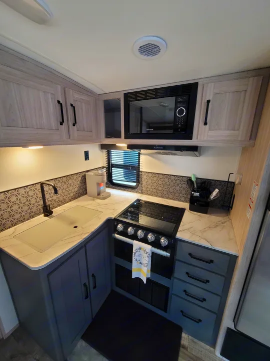 RV rental kitchen featuring light wood-look upper cabinets, dark gray lower cabinets, microwave, stove, sink, and modern backsplash.. Heartland RVs Sundance Ultra Lite 2022