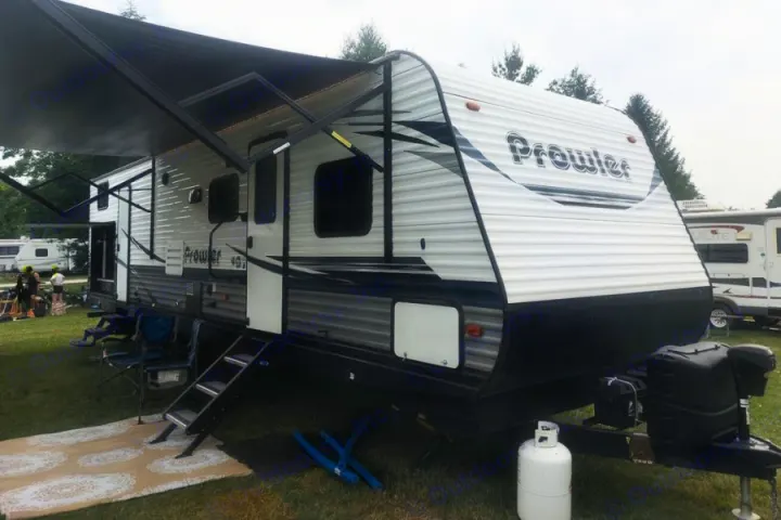 White & black Prowler travel trailer RV rental, awning extended over campsite setup with steps, rug, and chairs.. Heartland RVs Prowler 2020