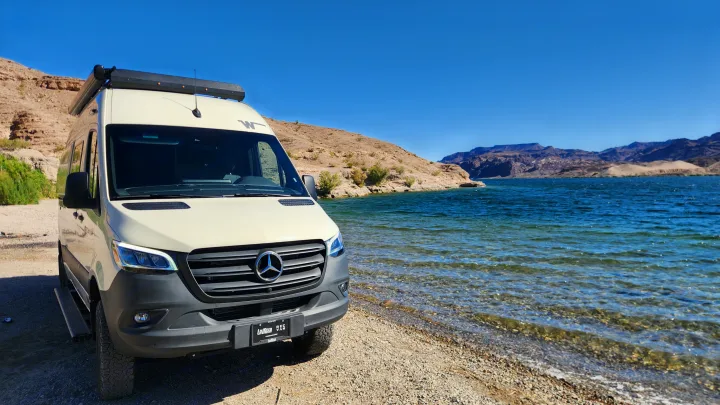 Cream Mercedes RV rental parked on a gravel shore next to a clear blue lake with mountains in the background.. Winnebago Revel 2024