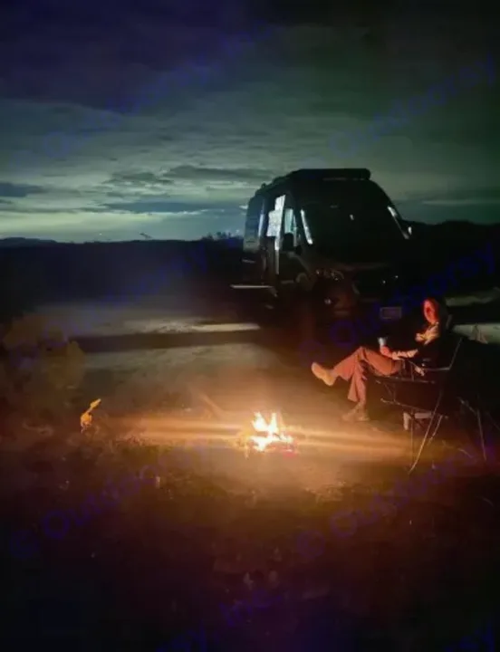 Nighttime RV rental adventure: Person relaxing by a warm campfire next to a camper van under a cloudy sky.. Winnebago Revel 2022