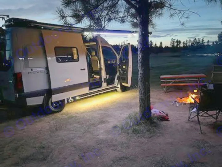 A white camper van from an RV rental, with its awning lit, parked beside a warm campfire at dusk.. Winnebago Revel 2022