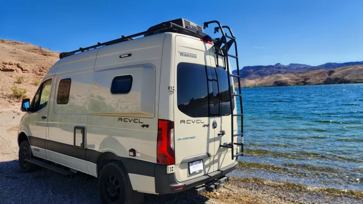 A cream-colored Winnebago Revel 4x4 rv rental camper van parked by a bright blue lake with mountains in the distance. Perfect for outdoor adventure.. Winnebago Revel 2022