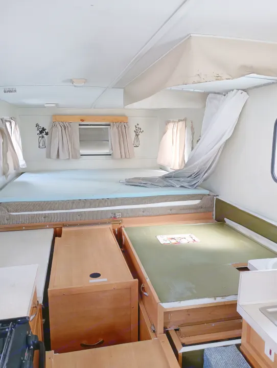 Inside an RV rental: view of light blue mattresses on sleeping platforms, kitchen counter, and wooden storage in a compact camper.. Trailmanor Trailmanor Trailer 2005