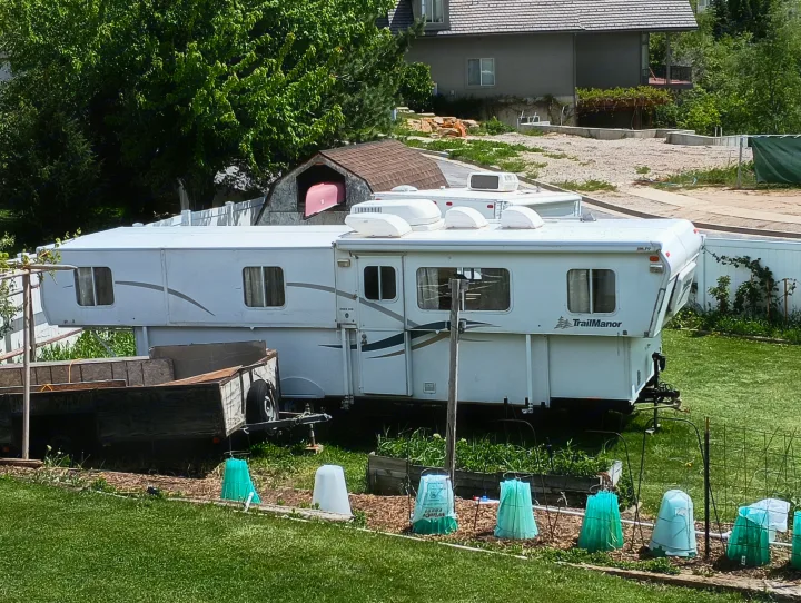 White TrailManor RV/travel trailer parked in a yard with a garden, small trailer, and a red boat. Great for an RV rental.. Trailmanor Trailmanor Trailer 2005