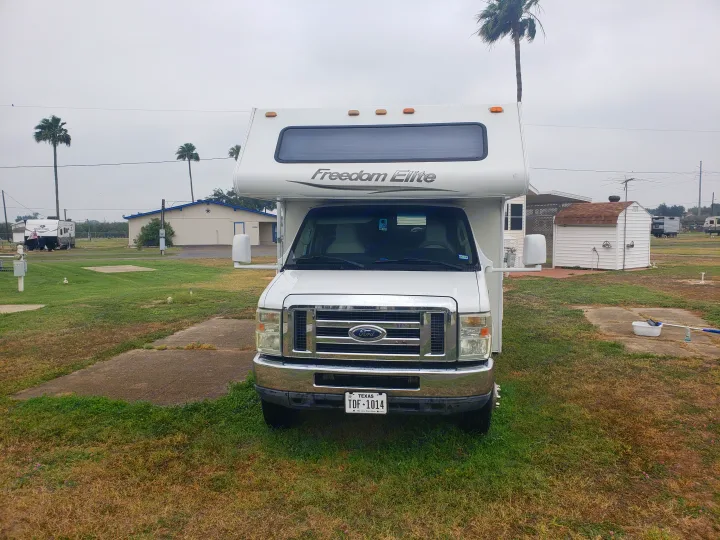 White Ford Freedom Elite RV rental parked at a Texas RV park, showing its front, surrounding grass, and distant buildings under an overcast sky.. Four Winds Freedom Elite 2010