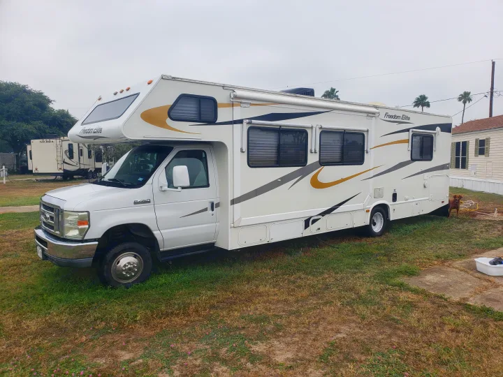 White Freedom Elite Class C RV rental on grass, with another RV & a dog nearby. Perfect for outdoor travel.. Four Winds Freedom Elite 2010
