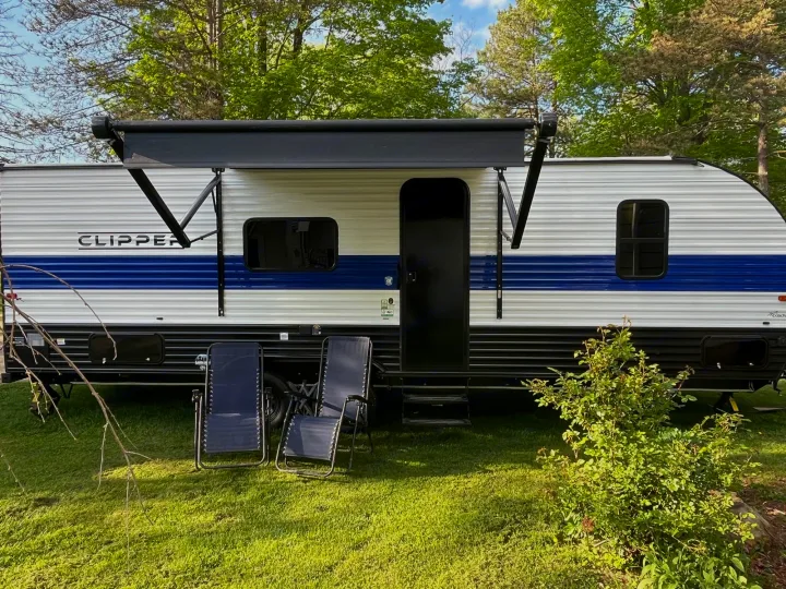 This blue and white RV rental travel trailer is set up for camping, with an extended awning and two chairs on the grass.. Coachmen Clipper 2024