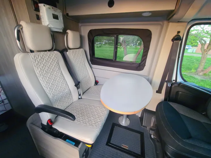 Comfortable interior of an RV rental camper van, showing passenger seats, a round dining table, and a scenic window view.. Winnebago Solis 2021