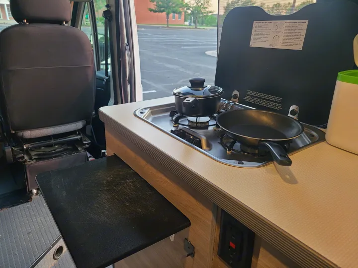 An RV rental's compact kitchen with a two-burner stove, pot, and pan, ready for cooking, next to a passenger seat.. Winnebago Solis 2021