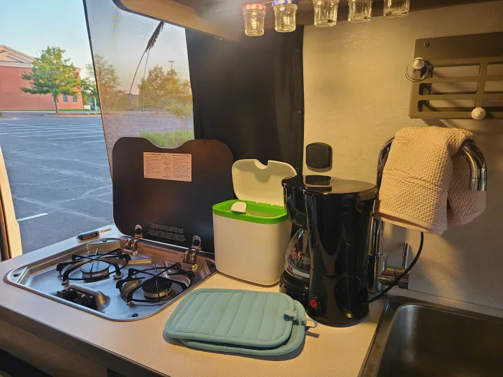 RV rental kitchen showing a stove, sink, coffee maker, and jars, with a window view of an outdoor parking lot.. Winnebago Solis 2021