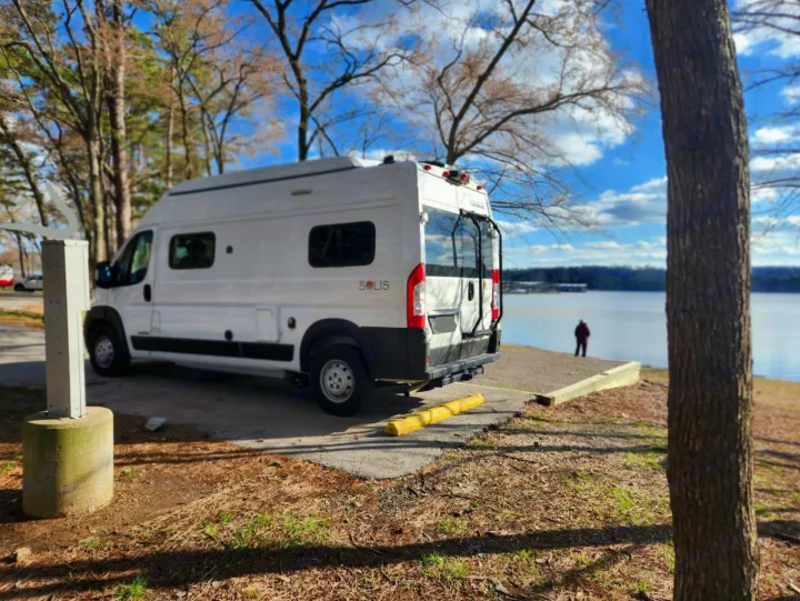White camper van RV rental parked at a serene lakeside spot, with a person enjoying the view. Ideal for outdoor adventures.. Winnebago Solis 2021