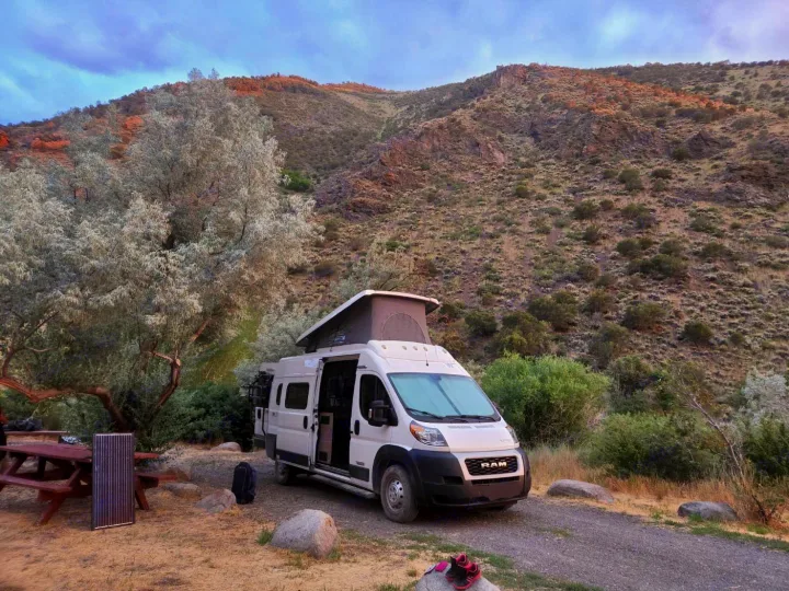 White RV rental camper van with pop-top parked at a scenic mountain campsite, next to a picnic table and solar panel.. Winnebago Solis 2021