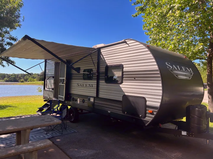 Serene lakeside RV rental: a "Salem" travel trailer with an extended awning, picnic table, and chairs on a sunny day.. Forest River Salem 2020