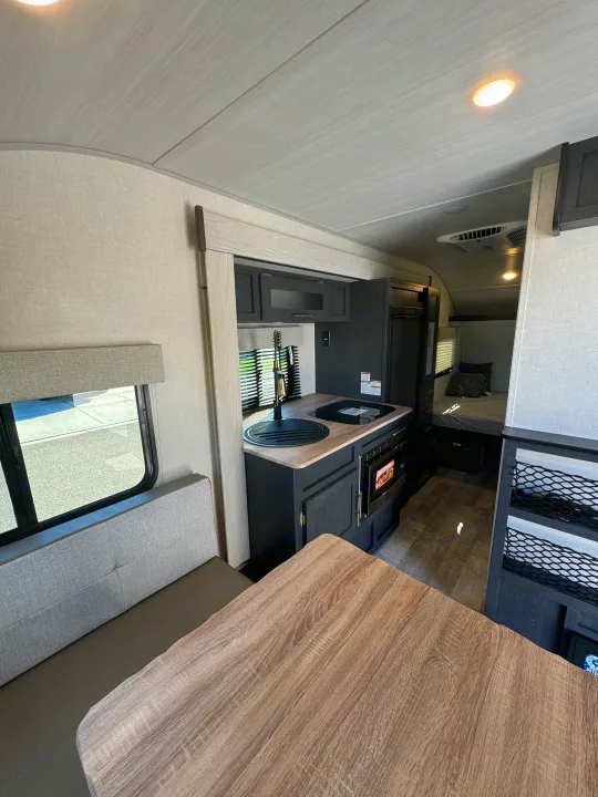View of an rv rental's modern interior: light wood-grain table, gray dinette, dark kitchen with sink, and a bed beyond.. R-Pod R-Pod Trailer 2024