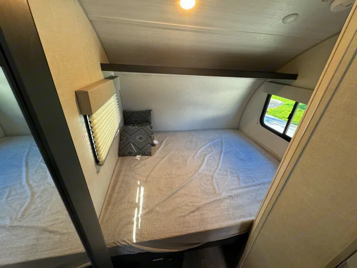 This inviting RV rental bedroom features a cozy bed with neutral linens, patterned pillows, a window, and overhead beam storage.. R-Pod R-Pod Trailer 2024