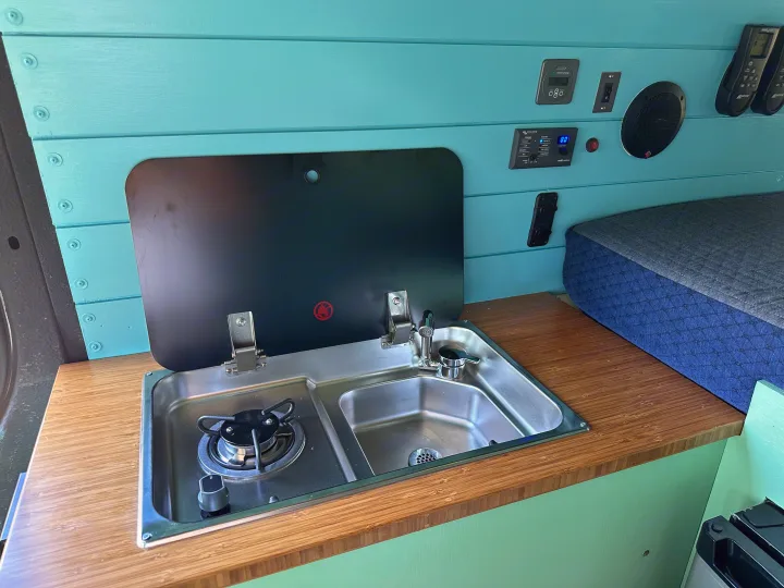RV rental interior featuring a teal kitchenette with stove and sink, plus essential power controls and a bed.. Mercedes-Benz Sprinter 2021