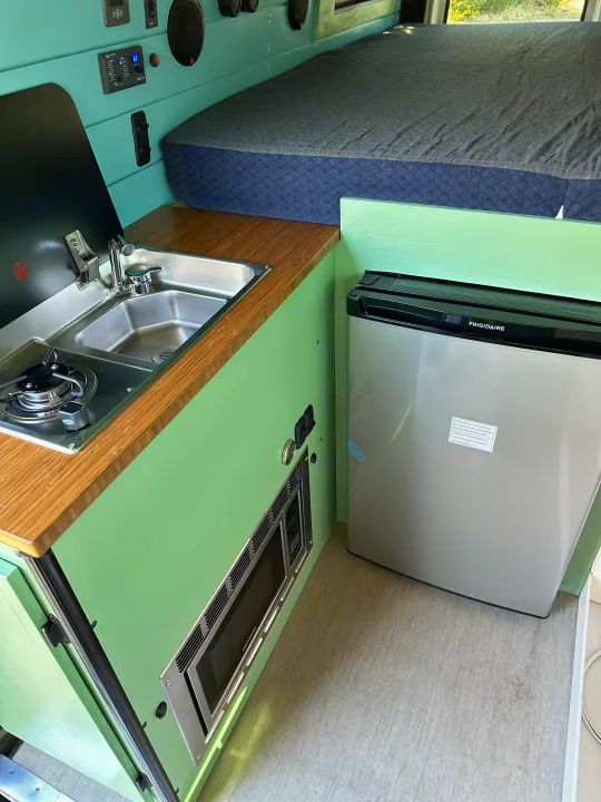 RV rental interior showcasing a modern kitchen with stove, sink, and microwave, plus a compact fridge and cozy bed.. Mercedes-Benz Sprinter 2021