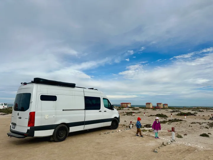 An RV rental van parked on a sandy beach with two kids exploring nearby small cabins under a cloudy sky.. Mercedes-Benz Sprinter 2021