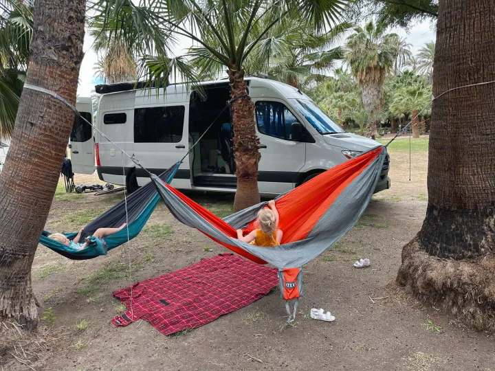 Kids relax in hammocks strung between palm trees at a serene camp spot, with their RV rental parked nearby.. Mercedes-Benz Sprinter 2021
