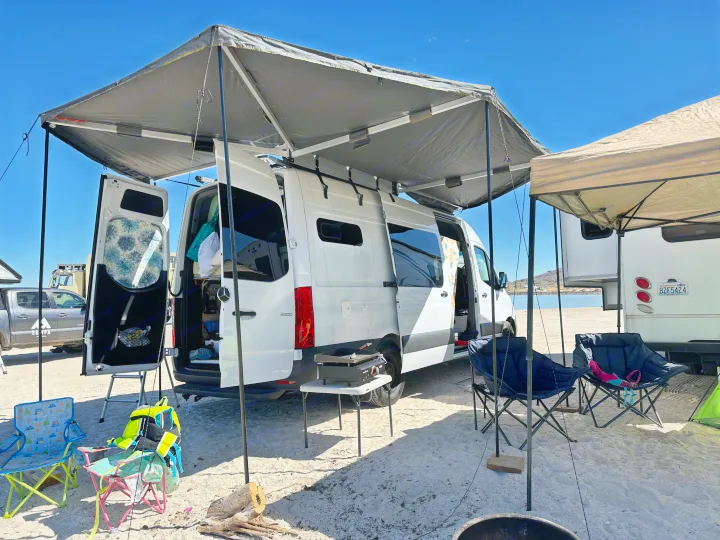 White RV rental camper van with awning, chairs, and grill set up on a sandy beach by the water, perfect for outdoor living.. Mercedes-Benz Sprinter 2021