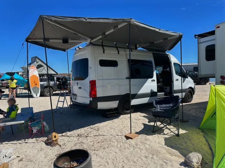 White camper van with an extended awning, set up at a sandy rv rental campsite. Perfect for sunny outdoor adventures.. Mercedes-Benz Sprinter 2021
