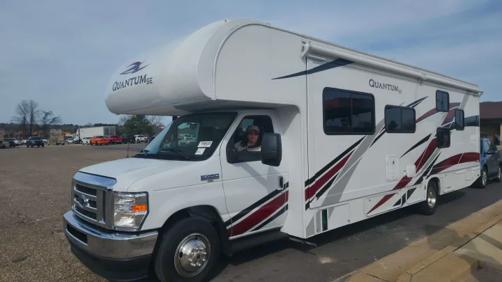Woman in a hat driving a white Class C Quantum SE rv rental with dark stripes, parked outdoors on a bright day.. Thor Quantum 2021