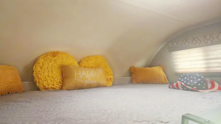 Cozy RV rental bedroom with a comfortable bed, cheerful yellow pillows, one reads 'HAPPY place,' and a patriotic hat.. Thor Quantum 2021