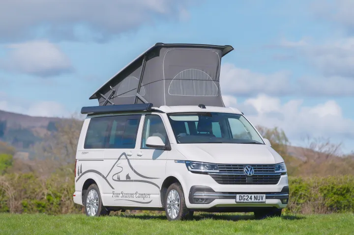 White Volkswagen campervan, likely an RV rental from Four Seasons Campers, with its pop-top roof up, parked in a sunny field.. Volkswagen California Ocean 2024