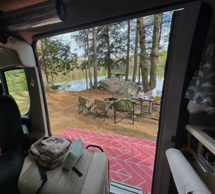 A cozy rv rental parked by a peaceful lake, showcasing a prepared campsite with chairs, a fire pit, and a picnic table for relaxing in nature.. Winnebago Travato 2015