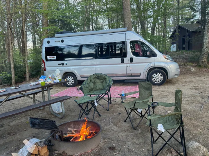Enjoying an RV rental in a wooded campsite with a roaring campfire, picnic table, and relaxing chairs. Perfect nature escape.. Winnebago Travato 2015